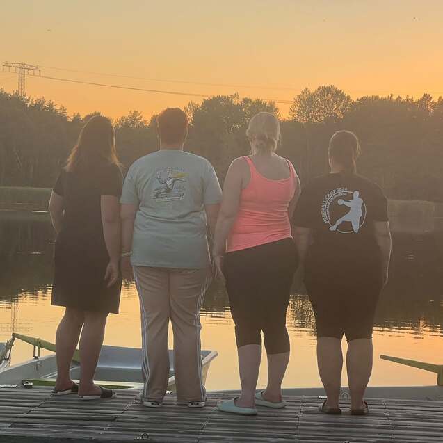 Vier Frauen stehen mit dem Rücken zu einem ruhigen See bei Sonnenuntergang auf einer Holzplattform.