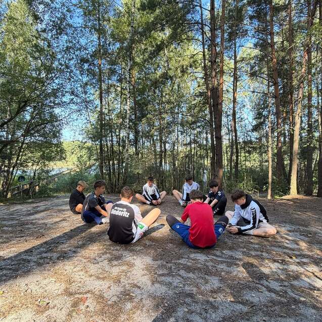 Gruppe von Jugendlichen sitzt im Kreis auf dem Boden im Wald, umgeben von hohen Bäumen und natürlichem Licht.