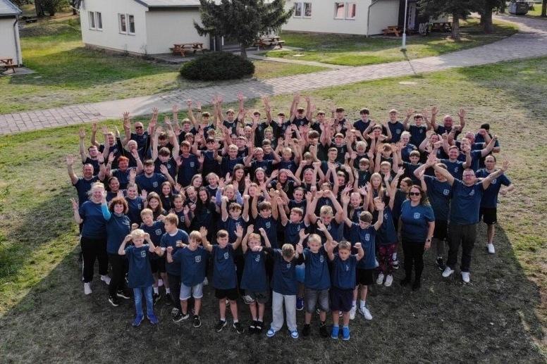 Gruppenfoto von &uuml;ber 150 Personen in blauen T-Shirts, die in einem Freiluftbereich jubeln. Im Hintergrund sind H&uuml;tten sichtbar.