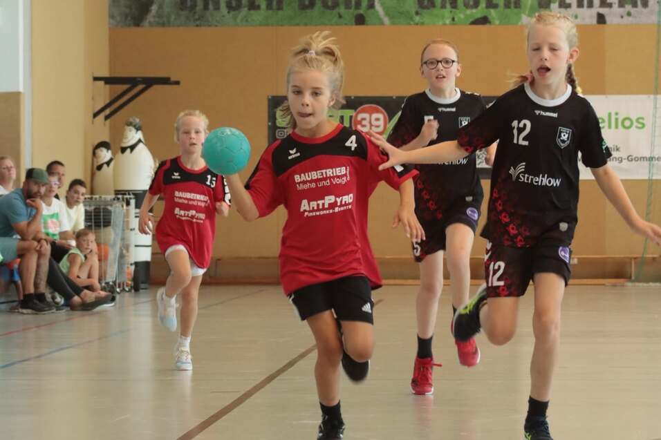 Vier Mädchen in Sportbekleidung spielen Handball in einer Sporthalle. Eine Spielerin mit Ball läuft voraus.