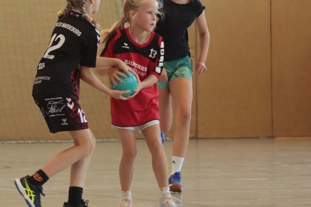 Drei Mädchen spielen Handball in einer Halle, eines hat einen Ball, während die anderen sich vorbereiten.