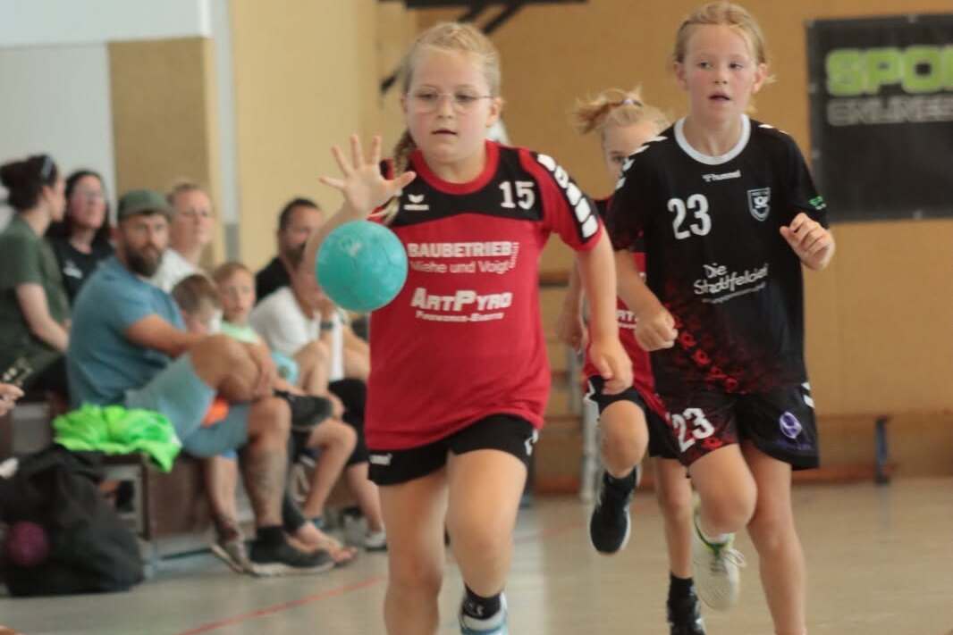 Zwei Mädchen spielen Handball in einer Sporthalle, eines dribbelt mit einem blauen Ball, während das andere folgt.