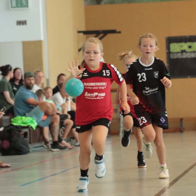 Zwei Mädchen spielen Handball in einer Sporthalle, eines dribbelt mit einem blauen Ball, während das andere folgt.