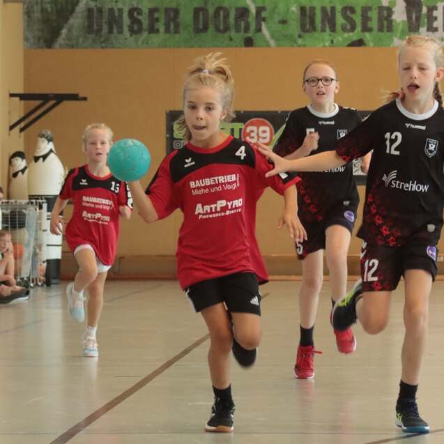 Vier Mädchen in Sportbekleidung spielen Handball in einer Sporthalle. Eine Spielerin mit Ball läuft voraus.