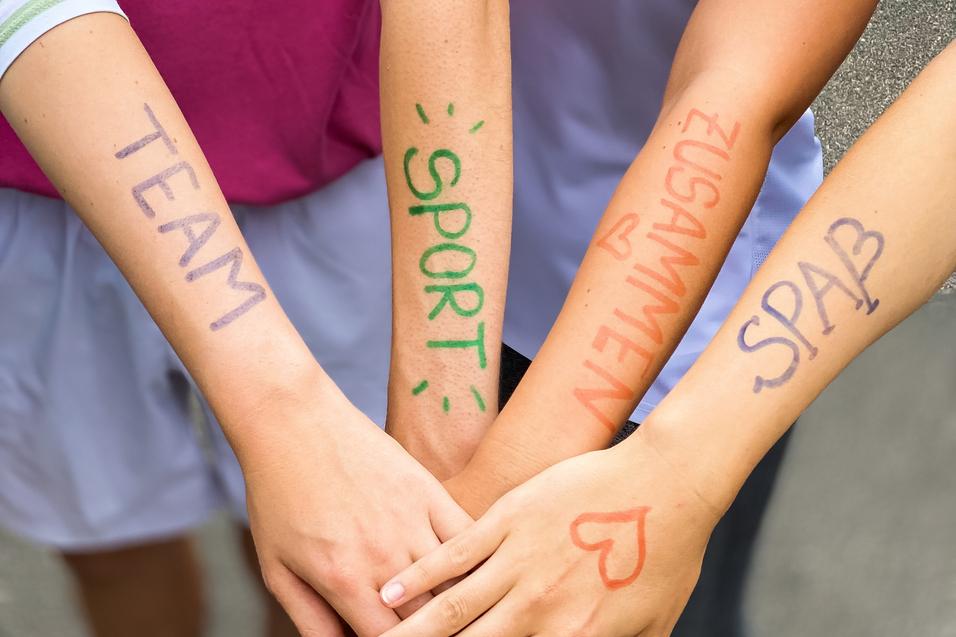 Vier Hände mit bunten Schriftzügen "TEAM", "SPORT", "ZUSAMMEN" und "SPASS" liegen übereinander auf einem grauen Untergrund.