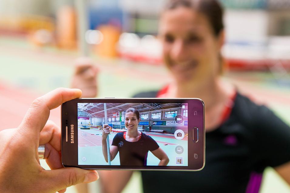 Hand hält ein Smartphone, das ein Porträt einer lächelnden Athletin mit Stab in einer Sporthalle zeigt.